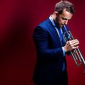 Ein junger Mann in einem eleganten blauen Anzug hält eine Trompete und spielt sie vor einem tiefrotem Hintergrund. Sein Blick ist nach unten gerichtet, was dem Bild eine konzentrierte und stimmungsvolle Atmosphäre verleiht.
