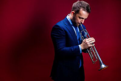Ein junger Mann in einem eleganten blauen Anzug hält eine Trompete und spielt sie vor einem tiefrotem Hintergrund. Sein Blick ist nach unten gerichtet, was dem Bild eine konzentrierte und stimmungsvolle Atmosphäre verleiht.