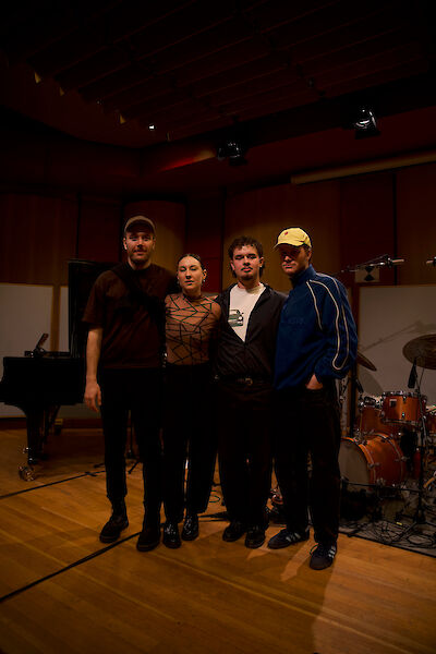 Vier Personen – drei Männer und eine Frau – stehen nebeneinander in einem professionellen Musikstudio und blicken in die Kamera. Im Hintergrund sind ein Flügel sowie ein Schlagzeug zu sehen, was auf eine Aufnahmesession oder ein Bandprojekt hindeutet.