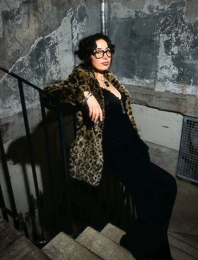 Eine junge Frau mit Brille sitzt auf einer Treppe in einem urbanen, industriellen Ambiente und trägt ein schwarzes Outfit kombiniert mit einem Leopardenmuster-Mantel und einer passenden Tasche. Das Foto hat einen stilvollen, modischen Charakter und wird durch das dramatische Lichtspiel vor der verwitterten Betonwand unterstrichen.