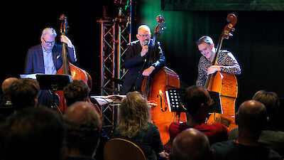 Drei Musiker stehen auf einer Bühne, wobei der mittlere ins Mikrofon spricht oder singt, während die beiden anderen jeweils einen Kontrabass spielen. Vor der Bühne sitzt ein aufmerksames Publikum, das der Veranstaltung in einem stimmungsvoll beleuchteten Konzertraum beiwohnt.