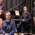 Vier Musikerinnen und Musiker posieren gemeinsam auf einer Bühne, im Hintergrund sind ein Kontrabass und ein Steinway-Flügel mit aufgelegten Notenblättern zu sehen. Die Gruppe besteht aus drei Männern und einer Frau, die locker und entspannt in die Kamera blicken.