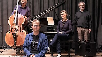 Vier Musikerinnen und Musiker posieren gemeinsam auf einer Bühne, im Hintergrund sind ein Kontrabass und ein Steinway-Flügel mit aufgelegten Notenblättern zu sehen. Die Gruppe besteht aus drei Männern und einer Frau, die locker und entspannt in die Kamera blicken.