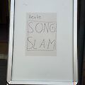 Ein weißes Aufstellschild mit Aluminiumrahmen steht vor einem Eingang. Darauf ist handgeschrieben auf einem aufgeklebten Zettel zu lesen: „Heute: Song Slam".