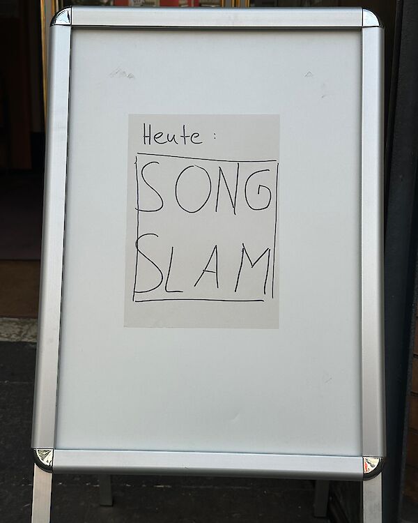 Ein weißes Aufstellschild mit Aluminiumrahmen steht vor einem Eingang. Darauf ist handgeschrieben auf einem aufgeklebten Zettel zu lesen: „Heute: Song Slam".