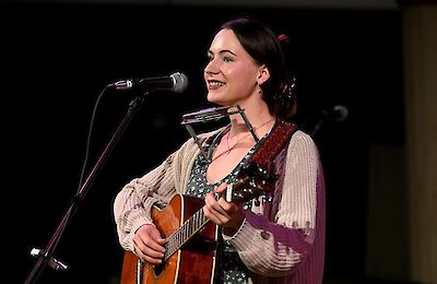Eine junge Frau mit dunklen Haaren steht lächelnd vor einem Mikrofon auf einer Bühne und spielt Akustikgitarre. Sie trägt einen gemusterten Träger-Overall, einen hellen Strickjacke und eine Mundharmonika-Halterung um den Hals.