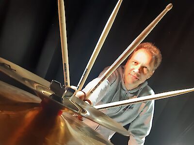 A man looks into the camera from behind a drum kit and several drumsticks, filmed from an unusual low-angle shot. The dramatic close-up shows the Rohema drumsticks in the foreground, whilst the drummer looks into the camera in the background.
