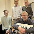 Vier Männer posieren gemeinsam für ein Gruppenfoto in einem schallgedämmten Raum, vermutlich einem Tonstudio oder Probenraum. Im Vordergrund lehnt einer der Männer auf einem Akkordeon, während die anderen drei lächelnd hinter ihm stehen.