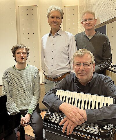 Four men are posing together for a group photo in a soundproof room, presumably a recording studio or rehearsal room. In the foreground, one of the men is leaning on an accordion, whilst the other three are standing behind him, smiling.