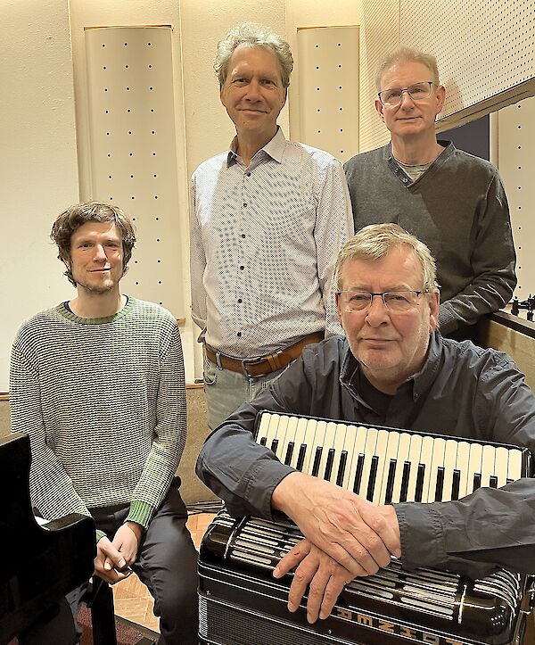 Vier Männer posieren gemeinsam für ein Gruppenfoto in einem schallgedämmten Raum, vermutlich einem Tonstudio oder Probenraum. Im Vordergrund lehnt einer der Männer auf einem Akkordeon, während die anderen drei lächelnd hinter ihm stehen.