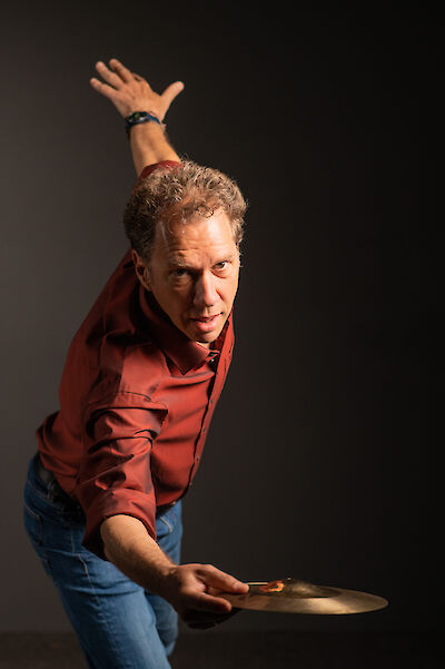 A man in a red shirt and jeans poses dynamically against a dark background, holding a cymbal in one hand and raising his other hand upwards. His determined gaze into the camera and his energetic posture convey the passion of a drummer.