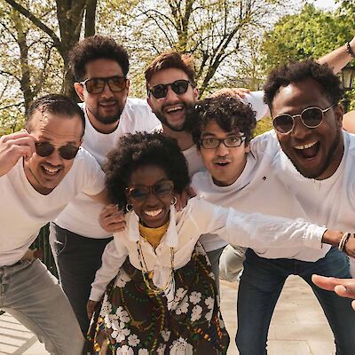 A group of six cheerful people are posing and laughing for a group photo outdoors, surrounded by trees in a sunny park setting. Most of them are wearing white T-shirts and sunglasses, whilst a woman in the centre is wearing a colourful floral dress.