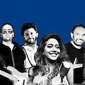 Eine Gruppe von sechs Musikerinnen und Musikern posiert vor einem blauen Hintergrund, wobei einige von ihnen Instrumente wie Gitarren und ein Saxofon halten. Das Bild ist in einem zweifarbigen Schwarz-Weiß-Stil gehalten, der dem Foto einen grafischen, plakatartigen Charakter verleiht.