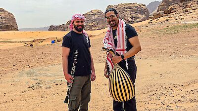 Two men are standing, smiling, in a desert landscape with imposing rock formations in the background. One is holding a clarinet and wearing a traditional Arabic headscarf (kufiya), whilst the other is holding an oud (Arabic lute) and wearing a red and white scarf round his neck.