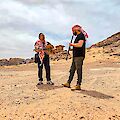 Zwei Männer musizieren gemeinsam inmitten einer kargen Wüstenlandschaft mit beeindruckenden Felsformationen im Hintergrund. Einer der Männer spielt eine Oud und trägt ein traditionelles arabisches Tuch, während der andere eine Klarinette spielt und ebenfalls ein rot-weißes Kufiya-Tuch trägt.