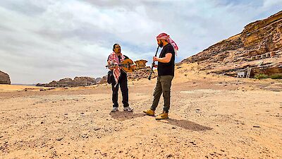 Two men are playing music together in the midst of a barren desert landscape, with impressive rock formations in the background. One of the men is playing an oud and is wearing a traditional Arab headscarf, whilst the other is playing a clarinet and is also wearing a red and white keffiyeh.