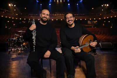 Two musicians are sitting side by side on a concert stage, smiling at the camera; in the background, the empty audience seats and stage lights of a concert hall can be seen. One is holding a clarinet, the other an oud; both are wearing black.