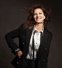 A smiling woman with long brown hair blowing in the wind poses confidently with one hand on her hip against a dark background. She is wearing a black blazer over a white shirt and exudes a lively, dynamic aura.