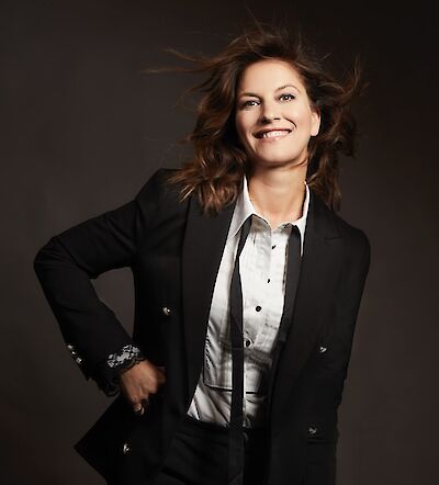 A smiling woman with long brown hair blowing in the wind poses confidently with one hand on her hip against a dark background. She is wearing a black blazer over a white shirt and exudes a lively, dynamic aura.