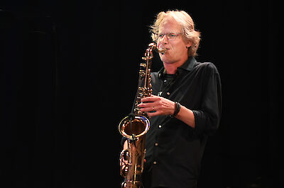 A middle-aged man with shoulder-length blond hair and glasses is playing a golden saxophone against a dark background. He is wearing a black shirt and appears to be on stage.