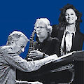 Drei Musiker posieren vor einem blauen Hintergrund: Ein Mann spielt Klavier, ein weiterer Mann spielt Saxofon, und eine lächelnde Frau steht im Hintergrund. Das schwarz-weiße Foto vermittelt eine elegante, jazzige Atmosphäre.