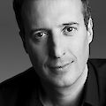 Das Foto ist schwarz-weiß. Es zeigt einen Mann. Der Mann ist nicht jung und nicht alt. Er hat kurze dunkle Haare. Er lächelt ein bisschen. Er trägt ein dunkles Hemd. Er schaut in die Kamera. Der Hintergrund ist grau.