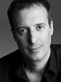 A black-and-white portrait photograph shows a middle-aged man with short dark hair and a slight smile. He is wearing a dark shirt and is looking directly at the camera against a plain grey background.