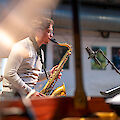 Ein junger Mann mit Brille spielt konzentriert ein goldenes Tenorsaxofon vor einem Mikrofon auf einer Bühne. Im Hintergrund sind Bilder an der Wand sowie weitere Musikinstrumente und Bühnenelemente zu erkennen.