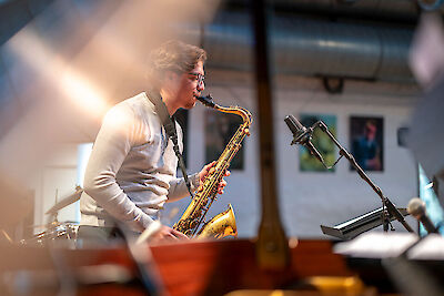A young man wearing glasses is intently playing a golden tenor saxophone in front of a microphone on stage. In the background, pictures on the wall, as well as other musical instruments and stage props, can be seen.