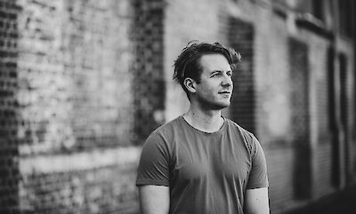 A young man with curly hair stands in front of a blurred brick wall in a black-and-white portrait photograph. He is wearing a plain T-shirt and is looking thoughtfully to one side.