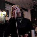 Eine junge Frau singt in ein Mikrofon. Sie hat lockiges Haar. Sie singt bei einem Konzert. Der Raum hat buntes Licht. Im Hintergrund sind Bilder an der Wand. Es gibt auch Pflanzen. Und noch eine weitere Person ist zu sehen.