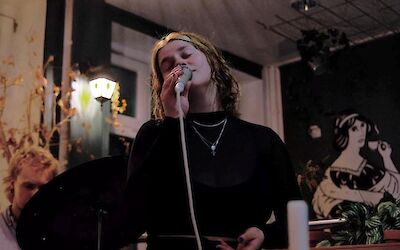 A young woman with curly hair is singing into a microphone during a live performance in an atmospherically lit room. In the background, there are decorative murals, plants and another person.