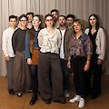 A group of about ten young adults are standing in front of a light grey curtain for a group photo. They are wearing a variety of outfits, ranging from casual to artistic and individual; one person in the middle is wearing striking red glasses.
