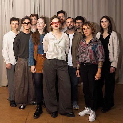 Eine Gruppe von etwa zehn jungen Erwachsenen steht für ein gemeinsames Gruppenfoto vor einem hellgrauen Vorhang. Die Personen tragen verschiedene lässige bis künstlerisch-individuelle Outfits, eine Person in der Mitte trägt auffällige rote Brillen.