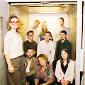 Eine Gruppe von etwa zehn jungen Erwachsenen posiert gemeinsam in und vor einem geöffneten Lastenaufzug oder Fahrstuhl. Das Foto hat einen experimentellen, leicht überbelichteten Look mit Bewegungsunschärfen im Hintergrund, was ihm eine künstlerische Ästhetik verleiht.