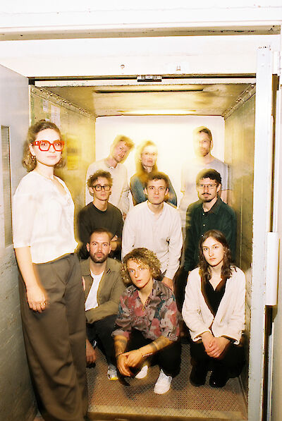 Eine Gruppe von etwa zehn jungen Erwachsenen posiert gemeinsam in und vor einem geöffneten Lastenaufzug oder Fahrstuhl. Das Foto hat einen experimentellen, leicht überbelichteten Look mit Bewegungsunschärfen im Hintergrund, was ihm eine künstlerische Ästhetik verleiht.