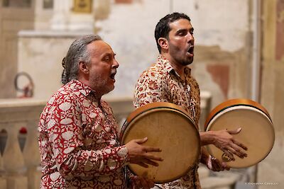 Zwei Männer in gemusterten Hemden spielen Rahmentrommeln und singen bei einem Auftritt in einem historischen Gebäude mit verputzten Steinwänden. Das Foto wurde von Giovanna Cavallo aufgenommen.