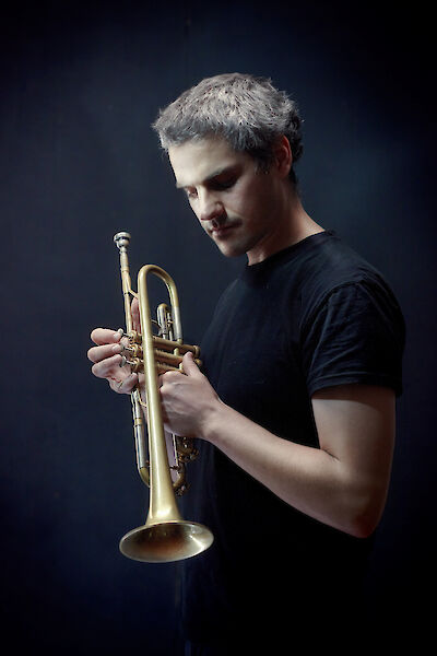 Ein Mann mit grauem Haar und schwarzem T-Shirt hält eine goldene Trompete in den Händen und blickt nachdenklich auf das Instrument herab. Das stimmungsvolle Studiofoto mit dunklem Hintergrund verleiht der Aufnahme eine künstlerische, melancholische Atmosphäre.