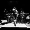 Ein Schwarz-Weiß-Foto zeigt einen Tänzer oder Performer von hinten auf einer Bühne, der eine dynamische Bewegung ausführt. Im Hintergrund sind zwei Musiker zu sehen – ein Schlagzeuger auf der linken und ein Keyboarder auf der rechten Seite –, die die Performance live begleiten.