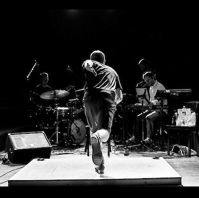 Ein Schwarz-Weiß-Foto zeigt einen Tänzer oder Performer von hinten auf einer Bühne, der eine dynamische Bewegung ausführt. Im Hintergrund sind zwei Musiker zu sehen – ein Schlagzeuger auf der linken und ein Keyboarder auf der rechten Seite –, die die Performance live begleiten.