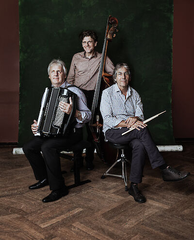 Three musicians are posing for a professional group photograph against a dark green background: one man is holding an accordion, another is sitting with drumsticks in his hands, and the third is standing in the middle with a double bass. The picture looks like a press photograph of a jazz or world music band.