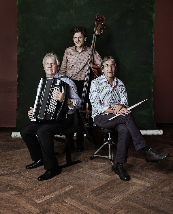 Drei Musiker posieren für ein professionelles Gruppenfoto vor einem dunkelgrünen Hintergrund: Ein Mann hält ein Akkordeon, ein anderer sitzt mit Drumsticks in der Hand, und der dritte steht mit einem Kontrabass in der Mitte. Das Bild wirkt wie ein Pressefoto einer Jazz- oder Weltmusik-Band.