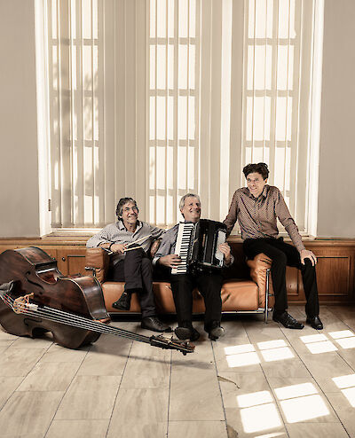 Three musicians are sitting, smiling, on a brown leather sofa in front of a bright window fitted with vertical blinds; the one in the middle is holding an accordion, whilst a double bass lies on the floor to the left. The warm, natural light and the relaxed atmosphere give the picture the feel of an atmospheric band photo.