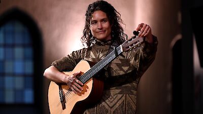 A person with long, curly hair is smiling whilst playing an acoustic guitar in an atmospherically lit room, with Gothic-style arched windows in the background. They are wearing a patterned jumper in shades of brown and gold and appear to be performing or recording.