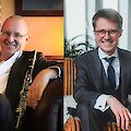Das Bild zeigt zwei Männer in eleganter Kleidung, die jeweils ein Musikinstrument halten – links ein Klarinettist mit einer Klarinette und rechts ein Trompeter mit einer Trompete. Beide lächeln freundlich in die Kamera und wirken wie professionelle Musiker, die für ein Porträtfoto posieren.