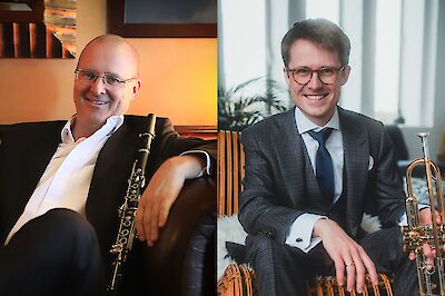 Zwei Männer stehen nebeneinander. Sie tragen elegante Kleidung. Der Mann links hält eine Klarinette. Der Mann rechts hält eine Trompete. Beide lächeln in die Kamera. Sie sind Musiker.