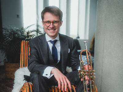 Ein Mann sitzt in einem hellen Raum. Er trägt einen Anzug und eine Krawatte. Er trägt auch eine Brille. Der Mann lächelt. Er hält eine Trompete in der Hand. Er sieht aus wie ein Musiker.