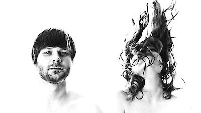 Ein schwarz-weißes Künstlerportrait zeigt zwei Personen vor weißem Hintergrund: links einen Mann mit kurzen Haaren und Dreitagebart, der ruhig in die Kamera blickt. Rechts ist eine Person zu sehen, deren langes, lockiges Haar dynamisch nach oben gewirbelt ist und das Gesicht verdeckt.