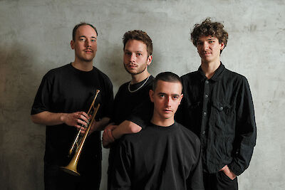 Vier junge Männer in schwarzer Kleidung posieren vor einer grauen Betonwand für ein stimmungsvolles Gruppenfoto. Der Mann auf der linken Seite hält eine goldene Trompete in der Hand, was auf ein professionelles Musikerporträt einer Band hindeutet.