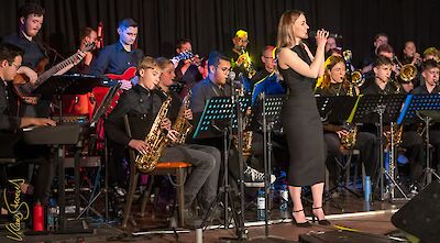 Eine junge Sängerin im schwarzen Kleid steht an einem Mikrofon und performt gemeinsam mit einer Big Band, die aus zahlreichen Musikern mit Saxofonen, Blechblasinstrumenten, Gitarren und weiteren Instrumenten besteht. Das Ensemble tritt bei einem Konzert auf einer professionell beleuchteten Bühne auf.